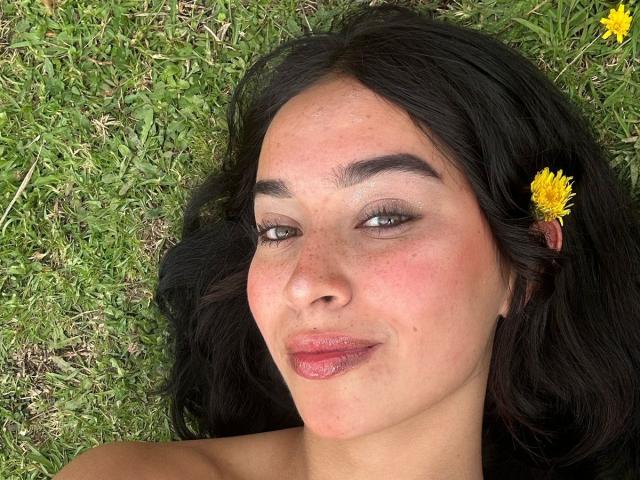 SolOrtega with dark wavy hair, a yellow daisy in her hair, and pinkish lips, lying on grass with a slight smile, showing freckles and light makeup.