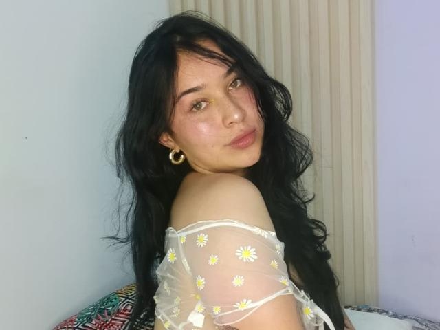 SolOrtega with long black hair, wearing a sheer white top with yellow daisy patterns, gold hoop earrings, and a small yellow dot on her nose, looking back.