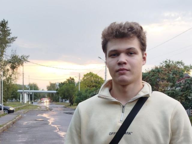 LucasLong has short brown curly hair, a neutral expression, and is wearing a beige zip-up hoodie with a black strap. He's standing on a wet, sunlit street.