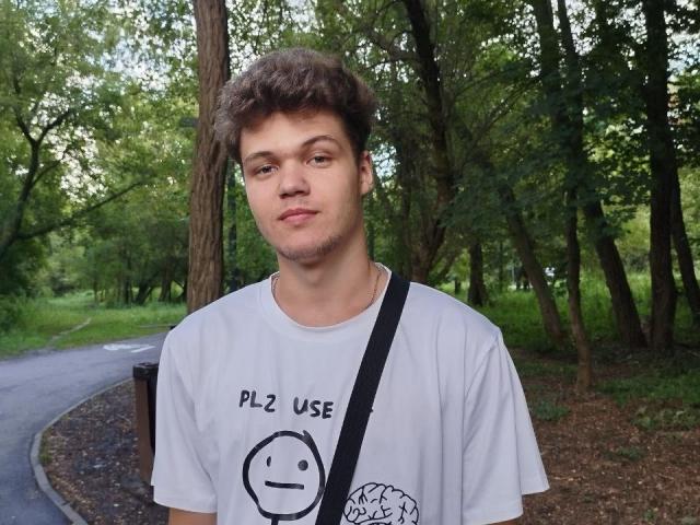 LucasLong with curly brown hair, wearing a white shirt with a smiley face and "PLZ USE" text, standing in a wooded area with a black strap across chest.