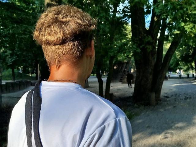 LucasLong with curly brown hair, wearing a white shirt and black backpack, facing away, standing in a sunlit, tree-filled park. His neck and upper back are visible.