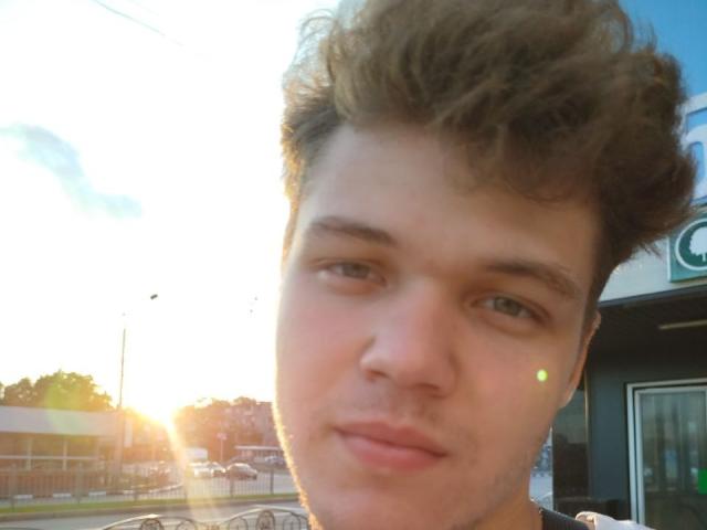 LucasLong with curly brown hair, light skin, green eyes, and a slight pout, sunlight flaring, looking off-camera. His nose has a small, visible freckle.