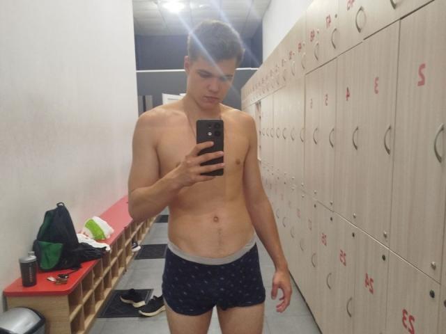LucasLong, shirtless, wearing dark blue boxer shorts with a pattern, holding a phone, taking a mirror selfie in a locker room, showing a toned, slim, and attractive upper body.