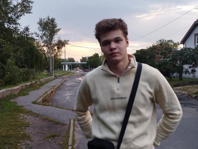 LucasLong with curly brown hair, wearing a beige "Chinamint" hoodie, black strap bag, standing on a wet, grassy, suburban street at sunset, hands on hips, looking at camera.