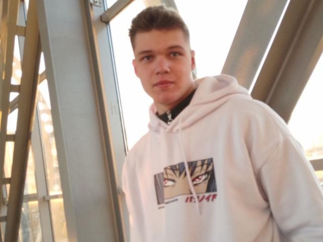 LucasLong with short brown hair, wearing a white hoodie with a graphic, standing next to metal beams, looking slightly to the side with a neutral expression.