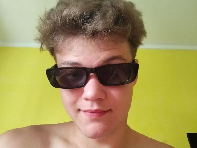 LucasLong with messy brown hair, wearing black sunglasses, smiling slightly, shirtless, against a yellow wall. His face is pale with light stubble.