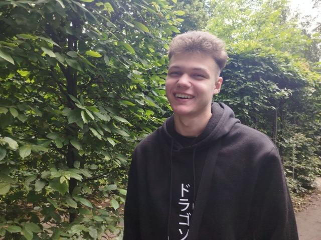 LucasLong, smiling with short brown hair, wearing a black hoodie, standing outdoors with green leafy background, visible from chest up, looking happy and relaxed.