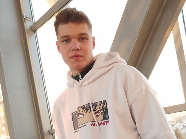 LucasLong, white hoodie with graphic, short brown hair, slight smirk, standing by window, metal frame in background, looking to camera, soft daylight, casual style, slightly leaning forward.