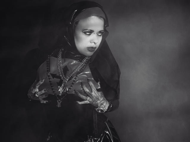 MorganaCrowly with dark hood, wearing latex gloves and dress, chains around neck, hands on tits, looking down, smoky black and grey photo, intense expression.