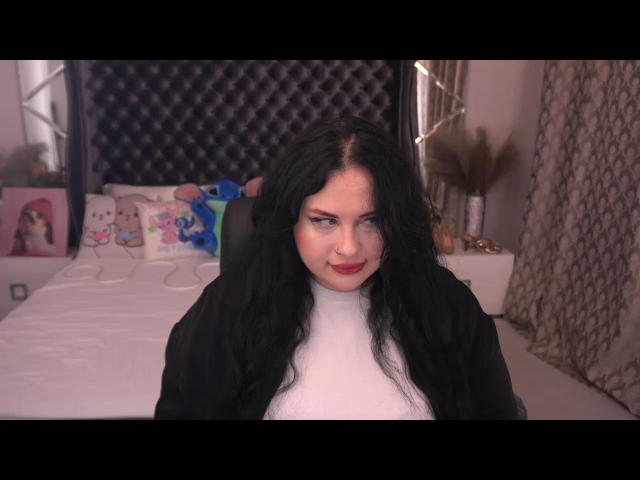 SerahMirabeli with long black hair, red lipstick, white turtleneck, and black jacket, sitting on bed, looking at camera with subtle smile. Boobs not visible. Bedroom setting.