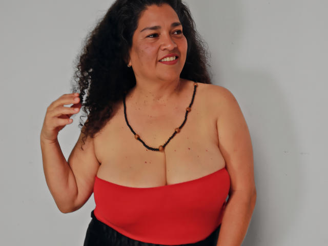 A curvy MaturexWoman with curly black hair, wearing a red strapless top, black necklace, and black pants, smiling at the camera, holding a cigarette.