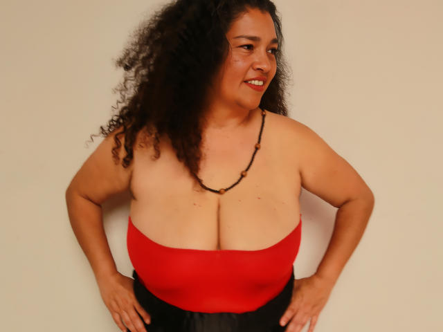 MaturexWoman with curly black hair, medium skin tone, and red strapless top showing large boobs, wearing black skirt and bead necklace, hands on hips, smiling slightly.