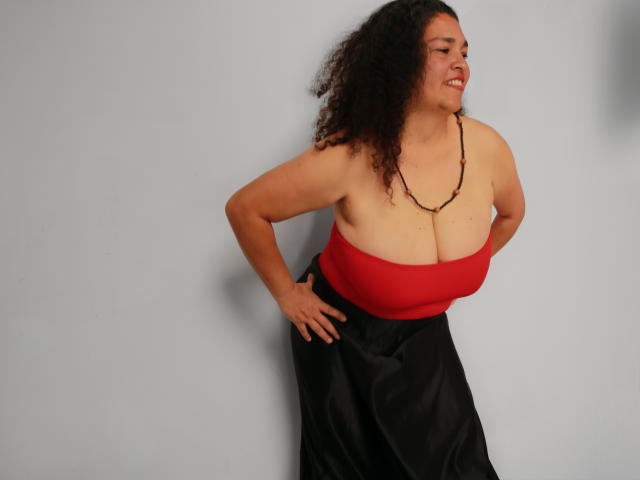 MaturexWoman with curly black hair, red strapless top showing big tits, black skirt, beaded necklace, hands on hips, smiling, standing against gray wall.