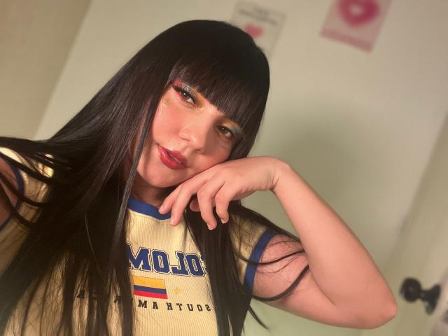 HollyKiss with long black hair, bangs, red lipstick, and yellow eyeshadow, wearing a yellow MOJO jersey, resting her chin on her hand, smiling softly.