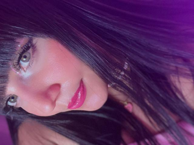 HollyKiss's face with pink lips, glossy pink lipstick, and black hair, her green eyes looking up with a slight smile, a soft pink blush on her cheeks.