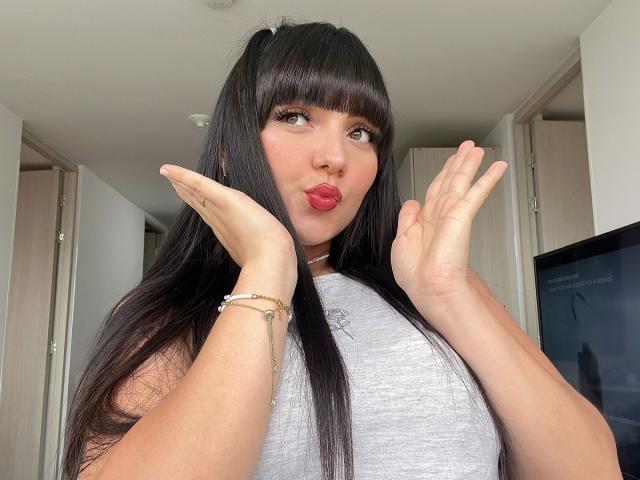 HollyKiss with long black hair, pouty red lips, and raised hands, wearing a gray sleeveless top, has a bracelet on her right wrist, in a bright room.