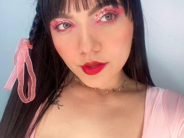 HollyKiss with pink eyeliner, red lipstick, black hair, pink ribbon, gold choker, pale skin, small tattoo on right shoulder, pink top, and a cute, sexy face.