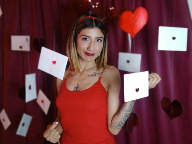 Kaiix with blonde hair, red dress, tattoos, holding white envelopes with red hearts, red headband, smiling, purple curtain, red heart balloon, and various heart-shaped decorations.