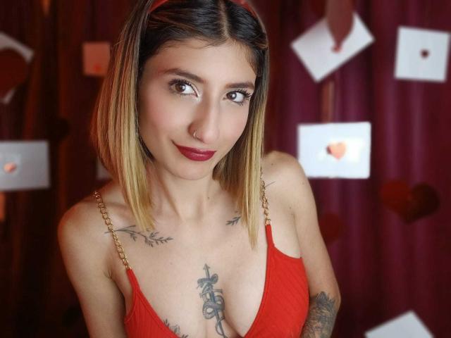 Kaiix, con cabello rubio hasta los hombros, vestido rojo, tatuajes en el pecho y el brazo, piercing en la nariz y lápiz labial rojo, sonriendo suavemente. Se ven los senos en el vestido rojo. Maquillaje ligero y colorete..