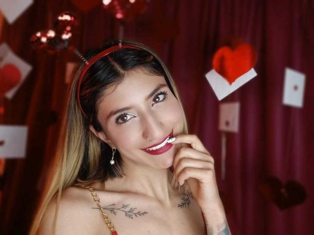 Kaiix, avec des cheveux blonds, un bandeau rouge, souriant, montrant ses dents, touchant son menton, des tatouages sur les clavicules, une chaîne en or, du rouge à lèvres et des boucles d'oreilles, avec des décorations en forme de cœur en arrière-plan..