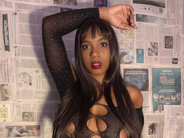 NataliKind, avec sa peau foncée, ses longs cheveux noirs et son rouge à lèvres rouge, porte un haut noir transparent à pois avec des découpes, bras levé, montrant ses seins et sa poitrine..