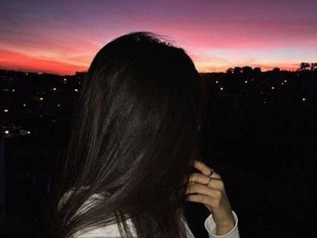 JolieBombonFontaine's dark brown, straight-ass hair covers her back, with a hand touching her head, in a white shirt, against a purple and pink sunset.