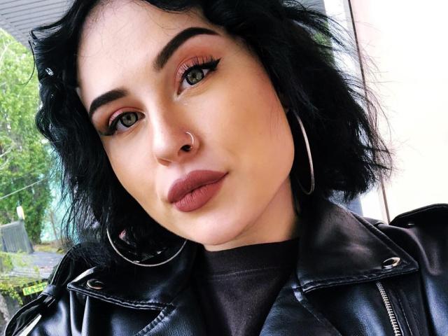 SerahMirabeli has black wavy hair, brown eyes, thick eyebrows, red eyeshadow, winged eyeliner, nose ring, and is wearing a black leather jacket with hoop earrings.