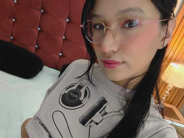 SophieDelaney, con gafas, lápiz labial rosa, cabello negro, suéter gris con auriculares y micrófono, sentada en la cama, cabecera tapizada de color rojo, pared de madera, lámpara en el fondo, mirando a la cámara..