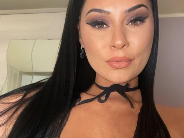 PrettyA with long black hair, dark eyeliner, and pink lipstick, wearing a black choker with a ribbon, has a slight smile and brown eyes, showing off her tits.