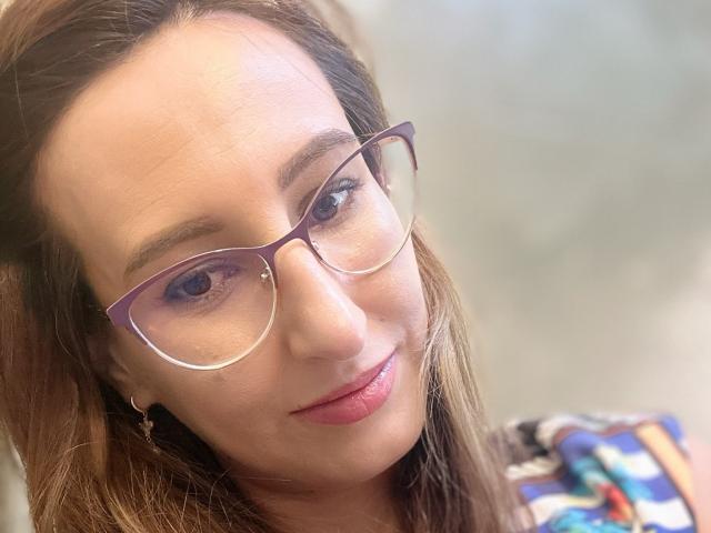 LysAddiction has light brown skin, blue eyes, and wears purple glasses with a small nose stud. Her brown hair is loose, and she's smiling with pink lipstick.