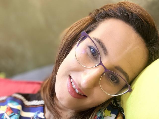 LysAddiction with purple glasses, brown hair, and a smile, laying on a green pillow, wearing a colorful patterned shirt, showing her cute face and white teeth.