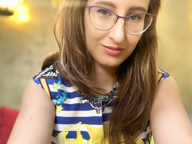 LysAddiction with glasses, brown hair, pink lips, wearing a blue and white striped dress with yellow star patterns, smiling slightly, in a bright room.