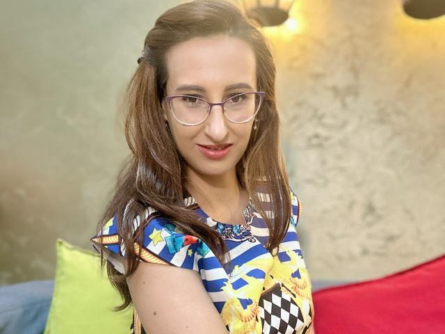 LysAddiction has brown hair, glasses, and a smile, wearing a colorful patterned dress, sitting with red and green pillows, and a textured wall behind her.