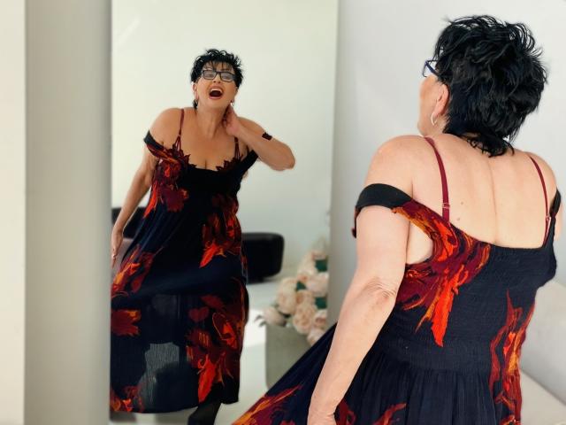 SondraRiddle with short black hair, glasses, and red lipstick, wearing a black dress with red floral patterns, standing in front of a mirror, looking at herself.