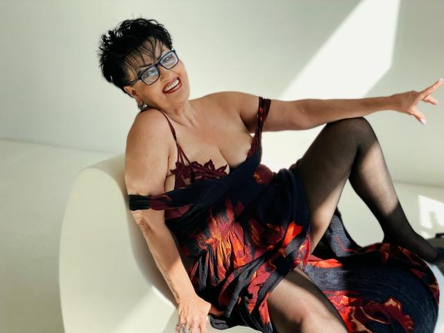 SondraRiddle with short black hair, glasses, and red lipstick, wearing a dark floral dress, black stockings, and off-shoulder, exposing her boobs, sitting on a white chair, smiling.