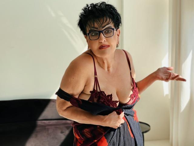 SondraRiddle with short black hair, glasses, and red lace bra, pulling down black and red lingerie to reveal her big, round boobs. She stands near a sunlit window.