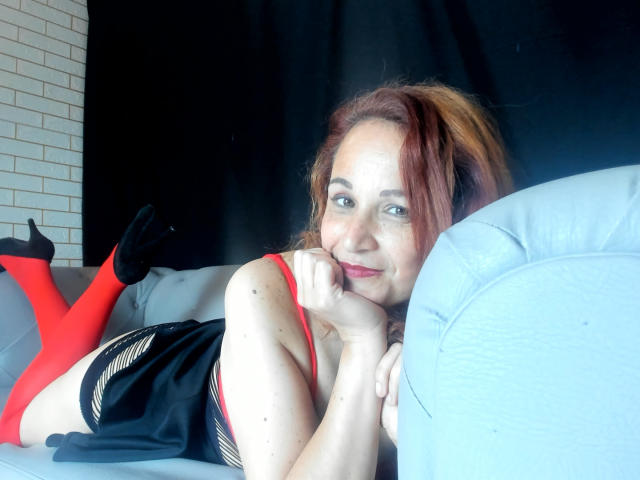 VictoriaLaRose lying on a couch, wearing a red top, black skirt, and red socks, with her hand on her chin, smiling, and her legs up, showing her ass.