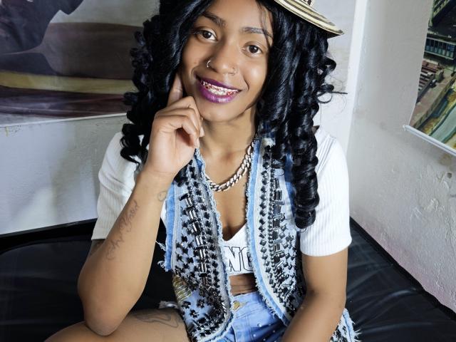 KatalinaCandy with dark brown skin, curly black hair, purple lipstick, white tee, blue patterned vest, gold necklace, smiling, nose ring, tattoo on right arm, sitting on black leather.