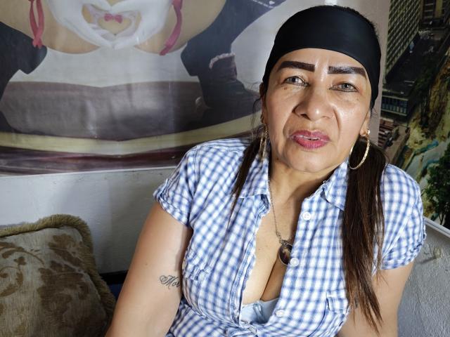 EstrellitaSexy, con ojos marrones, una diadema negra, aros grandes y una camisa a cuadros, mostrando parte de su escote y un tatuaje en el brazo, con una expresión ligeramente molesta. Se pueden ver sus senos..