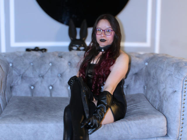 EvangelineJones with long dark red hair, black leather dress, black stockings, and gloves, sitting on a gray tufted couch, wearing a black collar and dark lipstick.