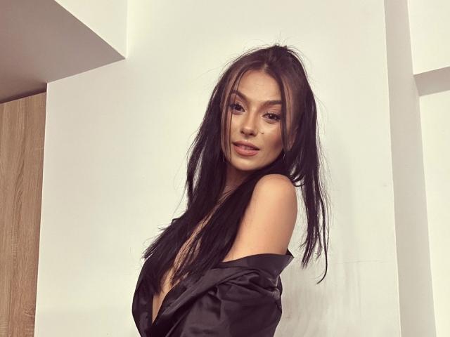 KinkyAmelie with long black hair, wearing a black off-shoulder shirt, exposing her tits, looking seductively at the camera, light skin, slight smile, soft lighting, indoor setting.