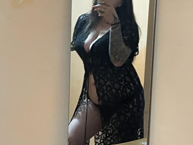 FortuneCarmen with tattoos, wearing a black lace robe, showing her tits and a slit revealing her pussy, taking a mirror selfie, covering her mouth with her hand.