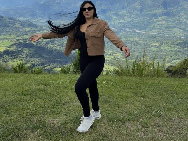 YessiJaymes with long black hair, wearing a brown jacket, black top, black leggings, and white sneakers, standing on grass with arms out, wearing sunglasses.