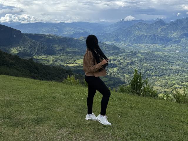 YessiJaymes with black hair, brown jacket, black pants, and white sneakers, standing on a grassy mountain, holding her hair, facing left, with a mountain landscape behind.
