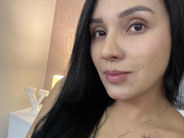 YessiJaymes with long black hair, brown eyes, nose ring, lip piercing, subtle makeup, and a slight pout, showing a gold necklace, in a soft-lit room.