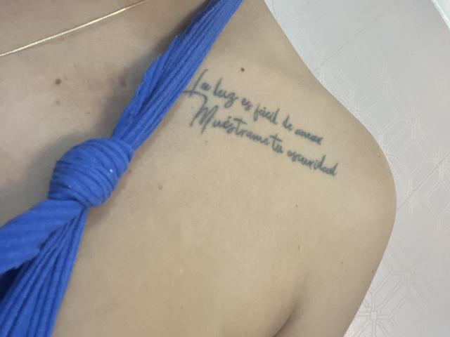 YessiJaymes' blue tied-up top shows a cursive tattoo on their shoulder reading "I love you more than words can say." YessiJaymes has light skin and a gold necklace.