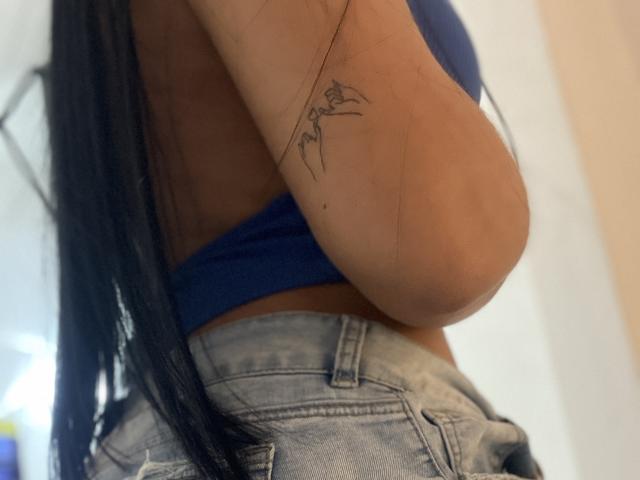 YessiJaymes has a small, black line tattoo on her upper arm, wearing a blue top and faded jeans, with long black hair. Her arm is raised.