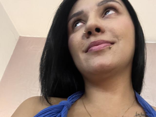 YessiJaymes with black hair, brown eyes, nose ring, lip piercing, blue halter top, looking up, smiling slightly, soft skin, subtle tattoo on left chest.