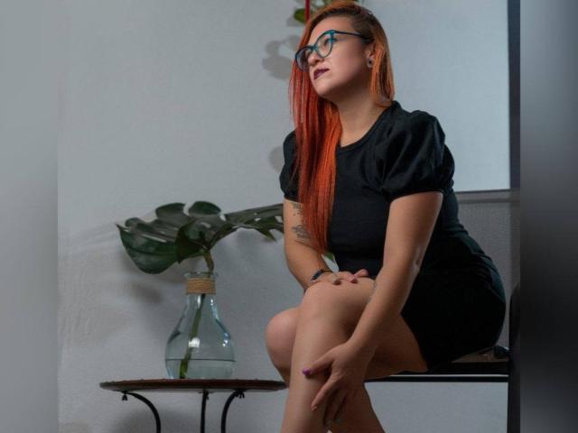 LunaRausch with red hair, glasses, black dress, sitting, legs crossed, showing her ass, tattoo on arm, potted plant, wooden table, white wall, looking to the side, serious.