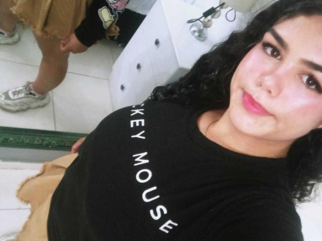 BrianaCleython with curly black hair, pink lipstick, and black t-shirt, showing her large tits in a mirror selfie. She's wearing beige shorts and white sneakers.